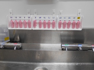ステンレス製のシンクの上部に、複数のピンク色の爪ブラシが整然と配置され、その下には手動操作部がない衛生的な水栓が二つ設置されている手洗い場のアップ写真