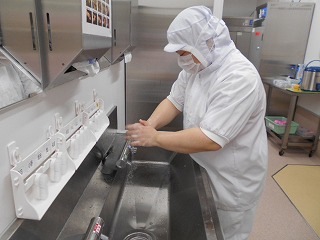 清潔なステンレス製の作業場で、白い帽子、マスク、作業着を着用した作業員が、シンクで両手を丁寧に洗っている写真