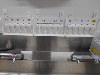 ステンレス製のシンクの上部に、複数の白色の爪ブラシが整然と配置され、その下には手動操作部がない衛生的な水栓が二つ設置されている手洗い場のアップ写真