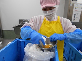 衛生的な白い作業服にピンクの衛生キャップ、黄色いエプロン、青い腕カバーと手袋を着用した作業員が、手で持ったボールに卵を割って、白い袋に卵の殻を入れている写真