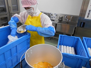 衛生的な白い作業服にピンクの衛生キャップ、黄色いエプロン、青い腕カバーと手袋を着用した作業員が、割った卵を小さなボウルから、ステンレス製の大きな容器に流し込んでいる写真