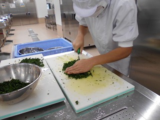 衛生的な白い作業服に白い衛生キャップとマスクを着用した作業員が、二枚の大きな白いまな板の上で、パセリのような濃い緑色の葉物野菜を、両手を使って細かくみじん切りにしている調理作業の写真