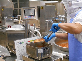 衛生的な白い作業服に白い衛生キャップとマスクを着用し、青い手袋をはめた作業員が、大きな柄杓で調理済みの料理をすくい上げ、計量器の上に置かれたステンレス製の容器で計量している写真