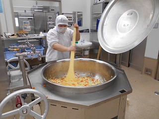 衛生的な白い作業服に白い衛生キャップとマスクを着用した作業員が、大型の回転釜に大量の細切りにされたキャベツとニンジンなどの野菜入れ、長い木べらを使って混ぜ合わせている調理風景の写真