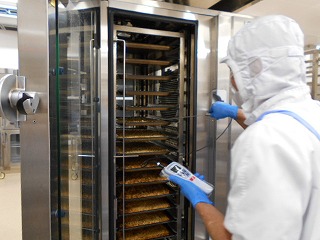 衛生的な白い作業服に白い衛生キャップを着用し、青い手袋をはめた作業員が、扉が開いた大型の業務用オーブンに近づき、食品に差し込んだ温度計の測定値を確認している写真