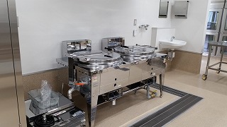 ピカピカのステンレス製のフラーヤーが2台並び、奥には手洗い場がある清潔感のある給食センターの写真