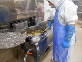 衛生的な白い作業服、白い衛生キャップ、青い手袋、青い腕カバー、青いエプロンを着用した作業員が、二槽の大型揚げ物用回転釜の片方で、泡立つ油の中を長い柄の調理器具で混ぜて調理している作業風景の写真