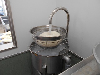 厨房内で、白米の入った大きなざるの下の銀色のバケツのような容器から湾曲した水道管が立ち上がり水が出て米を洗っている様子の写真