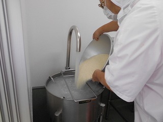 白衣の調理員が大きなステンレス製のボールに大量の米を入れ洗い、水を流している写真