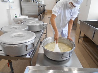 白衣とマスクを着けた調理員が、大きなステンレス製の円形容器に入った大量の米と具を手持ちのしゃもじでかき混ぜている写真