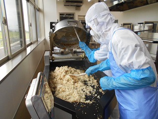 窓のある広い調理場で、白衣と青いエプロンを着けた二人の調理員が、自動回転釜で炊き上がった大量のご飯を巨大なしゃもじでかき混ぜ、混ぜご飯を作っている様子の写真