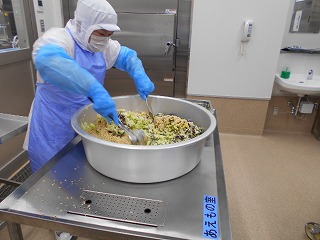 白衣と青いエプロンを着けた調理員が、大きな円形のアルミ製容器の中で、野菜などの具材を二本のしゃもじで混ぜ合わせている作業中の写真