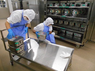 白衣とマスクを着けた二人の調理員が、青いエプロンと手袋を着用し、カートの上に積んでいる食缶をコンテナに積んでいる作業風景の写真