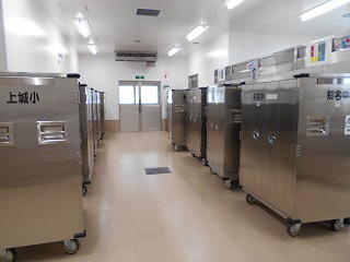 給食センターの清潔な通路に、学校名（上城小、知名中など）が書かれた、キャスター付きのステンレス製運搬用食缶カートが整然と並ぶ写真