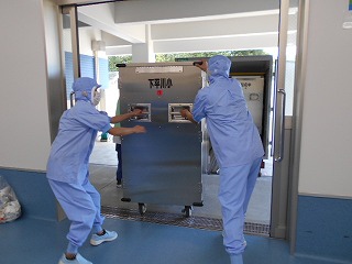 水色の防護服と帽子を着用した二人の調理員が、学校名が書かれた銀色の大型食缶運搬カートを両側から持って押し出し、建物の扉から運び出している写真