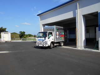 白い給食センターの建物から、かわいいイラストが側面に描かれた運搬用トラックが発車口からアスファルトの駐車場へ出ていく写真