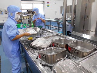 衛生的な作業服とマスク、帽子を着用した2名の作業員が、給食で使用された食缶をシンクで洗っている写真