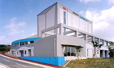 白い外壁を基調に灰色の壁面が組み合わさり、一部に水色のアクセントが入ったモダンな多目的施設で、正面には多くの窓があり、手前には舗装された道路と整備された芝生、その周囲に数本の小さな木が見えるあしびの郷・ちなの外観写真