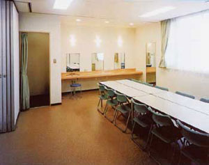 明るいベージュの壁の部屋で、右側には窓に沿ってメイクアップ用のライトのついた鏡が並び、手前には長机と椅子が並び、左奥のカーテンで仕切られた着替えスペースが一体となった楽屋内部の写真