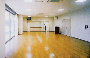 明るい木のフローリングの広い部屋で、左側に大きな窓、奥の壁には大きな鏡と手すりがあるリハーサル室内部の写真