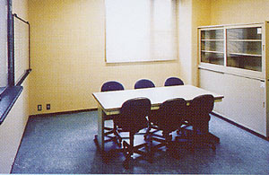 クリーム色の壁と濃い色の床の簡素な会議室のような部屋で、中央には四角いテーブルとキャスター付きのオフィスチェアが6脚あり、右奥にはガラス引き戸の収納棚、左側には窓とホワイトボードが設置されているスタッフ室内部の写真