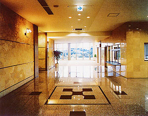 広々としたホールで、床中央には黒と茶色で幾何学模様の装飾が施され、奥には玄関の入り口が見えるエントランスホールの様子