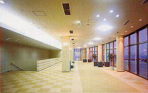 明るい色の四角い模様のカーペットが敷かれた広いロビーのような空間で、大きな窓が並び外の景色が見えるホワイエの写真