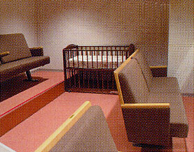 薄暗い室内で、ピンク色のカーペットが敷かれ、茶色の布張りのソファと中央には木製のベビーベッドが配置され、手前にも別のソファが置かれている親子室の写真