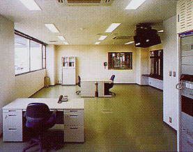 窓際の手前には引き出し付きの大きな机と黒いオフィスチェアがあり、その奥には別の机と椅子が配置され、壁には収納棚や大きなモニターも見える事務室内部の写真