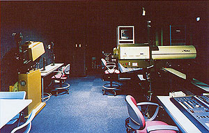 薄暗いスタジオの部屋で、青いカーペットが敷かれた床に、椅子や机、放送機材などの機器が整然と並びんでいる音響・照明調整室内部の写真