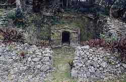 石灰岩の野面積みの低い石垣が中央に左右に伸び、中央の通路の奥、鬱蒼とした植物に覆われた斜面には石造りの小さな入り口が見えるアーニマガヤトゥール墓の写真