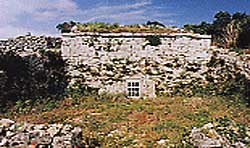 日当たりの良い空の下、白い岩の石垣に囲まれ、中央下部に小さな白色の窓を持つ石造りの建造物の屋者琉球式墳墓の写真