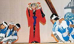 舞台の上で、頭には華やかな冠のような飾りをつけ赤い着物を着た女性が舞っており、両側に、青い衣装に白い鉢巻を締めた複数の男性しゃがんでいる大蛇踊りの写真