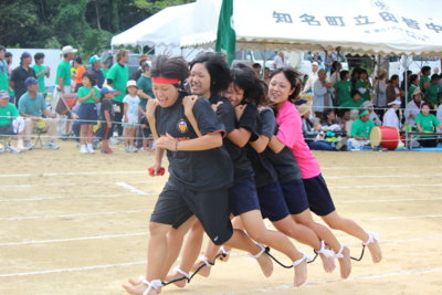 ムカデ競走で、5人の女性が肩を抱き合い、それぞれの足首を紐で繋いで一列になり、沿道の観客に見守られながら、懸命にゴールを目指して走っている様子の写真
