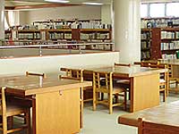 明るい光が差し込む図書館の室内で、手前には木製の閲覧机と椅子が整然と並んでおり、奥の壁際や柱の向こう側には多くの本が収納された背の高い木製の書棚が複数設置されている落ち着いた図書館内の様子を捉えた写真