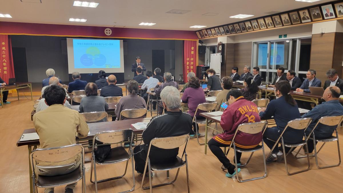 室内で開かれている住民説明会で、中央の壇上でスーツ姿の男性が参加者に向かってスクリーンに映し出された図を説明しており手前の木製の机とパイプ椅子に座った多くの参加者が熱心に耳を傾けている田皆字の説明会の写真