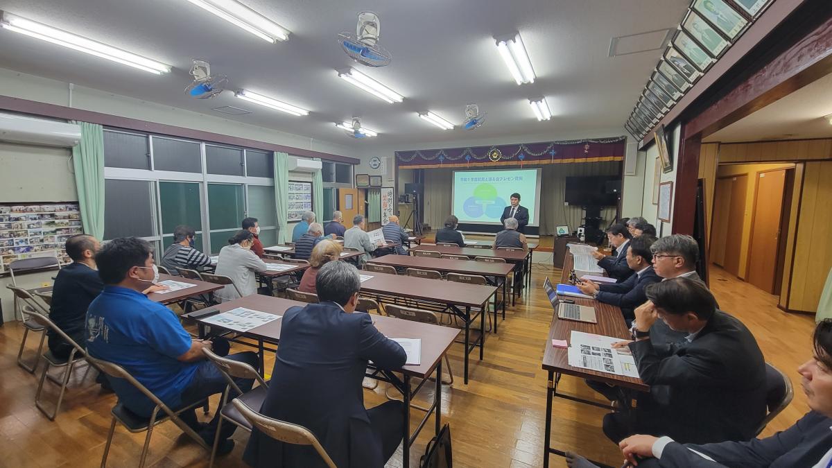 室内で壇上でスーツ姿の男性がスクリーンに映し出された資料を説明しており、会場に集まった数十人の聴衆が木製机とパイプ椅子に座って真剣に耳を傾け右側には関係者が座っている下城字の説明会の写真