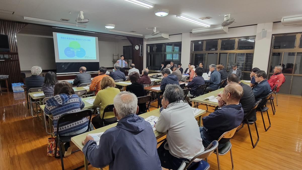 広々とした室内で、スーツ姿の男性が聴衆に向かってスクリーンに映し出された資料を説明しており、多くの参加者が机と椅子に座って真剣に耳を傾けている知名字の説明会の写真