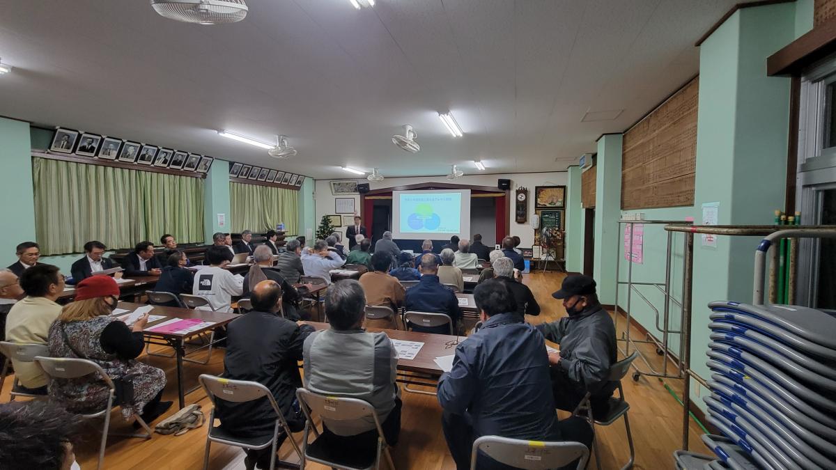 広々とした室内で、スーツ姿の男性が聴衆に向かってスクリーンに映し出された資料を説明しており、多くの参加者が机と椅子に座って真剣に耳を傾けている上平川字の説明会の写真