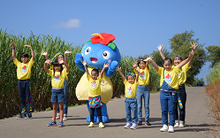 サトウキビ畑の間の道で青い体にハイビスカスを頭につけたマスコットキャラクターちなボーが、黄色いお揃いのTシャツと青いデニムを着用した8人の子どもたちと共に、両手を挙げてポーズを取っている写真