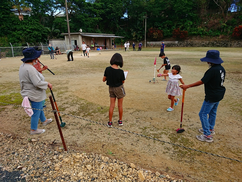 砂地の広場で複数の人々がスティックを使ってボールを打つグラウンドゴルフを楽しんでおり、中央にはスコアを記録する子どもとピンクの旗付きホールがある屋外運動場の写真。