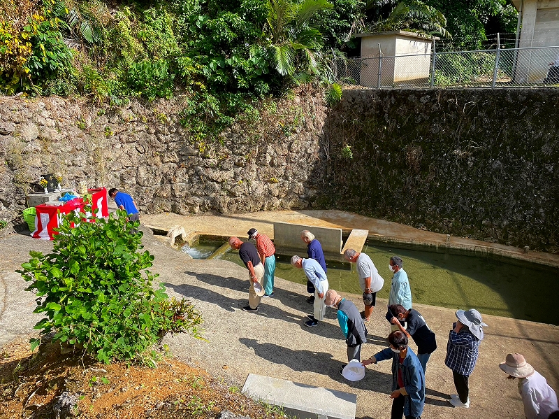 石垣に囲まれた湧水池の前で複数の人々が整列し、頭を下げて礼をしており、左奥には赤と白の布で覆われた祭壇が置かた水神祭の写真。