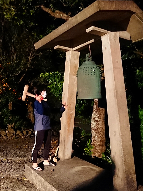 石製の柱で支えられた屋根の下に吊るされた大きな青銅製のジョセキの鐘があり、横には木槌を構えた人物が立つ夜の屋外風景が広がる写真。