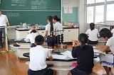 実験台が並ぶ理科室のような教室で、複数の生徒が実験に取り組んだり、教員から説明を聞いたりしている様子の写真