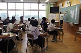教室の黒板の前で先生が生徒に説明をしており、多くの生徒が自席に座って授業を受けている様子の写真