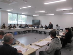 大きな窓のある会議室で、多くの参加者がロの字型に配置されたテーブルに着席し、一人の男性が立ち上がって説明をしている行政部会の様子の写真