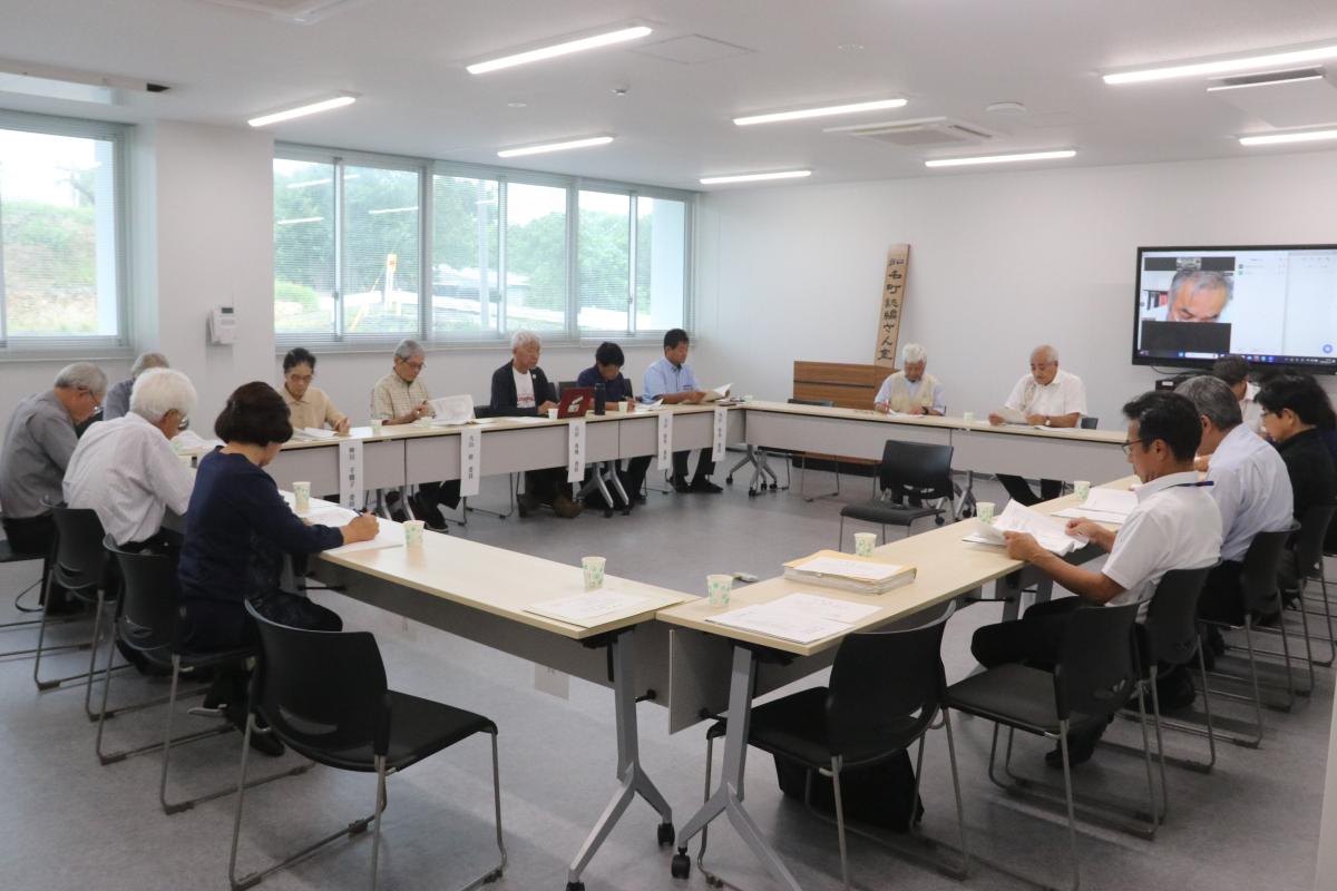 大きな窓のある部屋で、多くの参加者がロの字型に配置されたテーブルに着席し、前方のモニターに映る男性の話を聞いている編さん委員会の写真