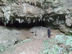 暗い洞窟の入り口が写っており、天井からは無数の鍾乳石が垂れ下がり、その手前には黒い服を着た男性が立っていて、周囲の遺跡の調査を行っている様子の写真