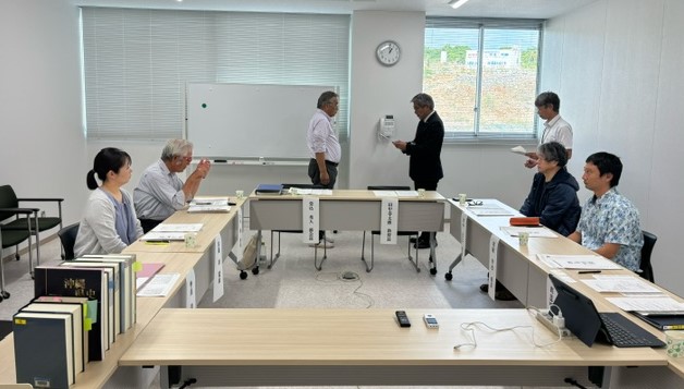 白い壁とブラインドがある会議室で、関係者が「コ」の字型に配置されたテーブルを囲んで会議を行っており、中央奥ではスーツ姿の男性が立ったまま資料のようなものを受け渡している考古部会の様子の写真