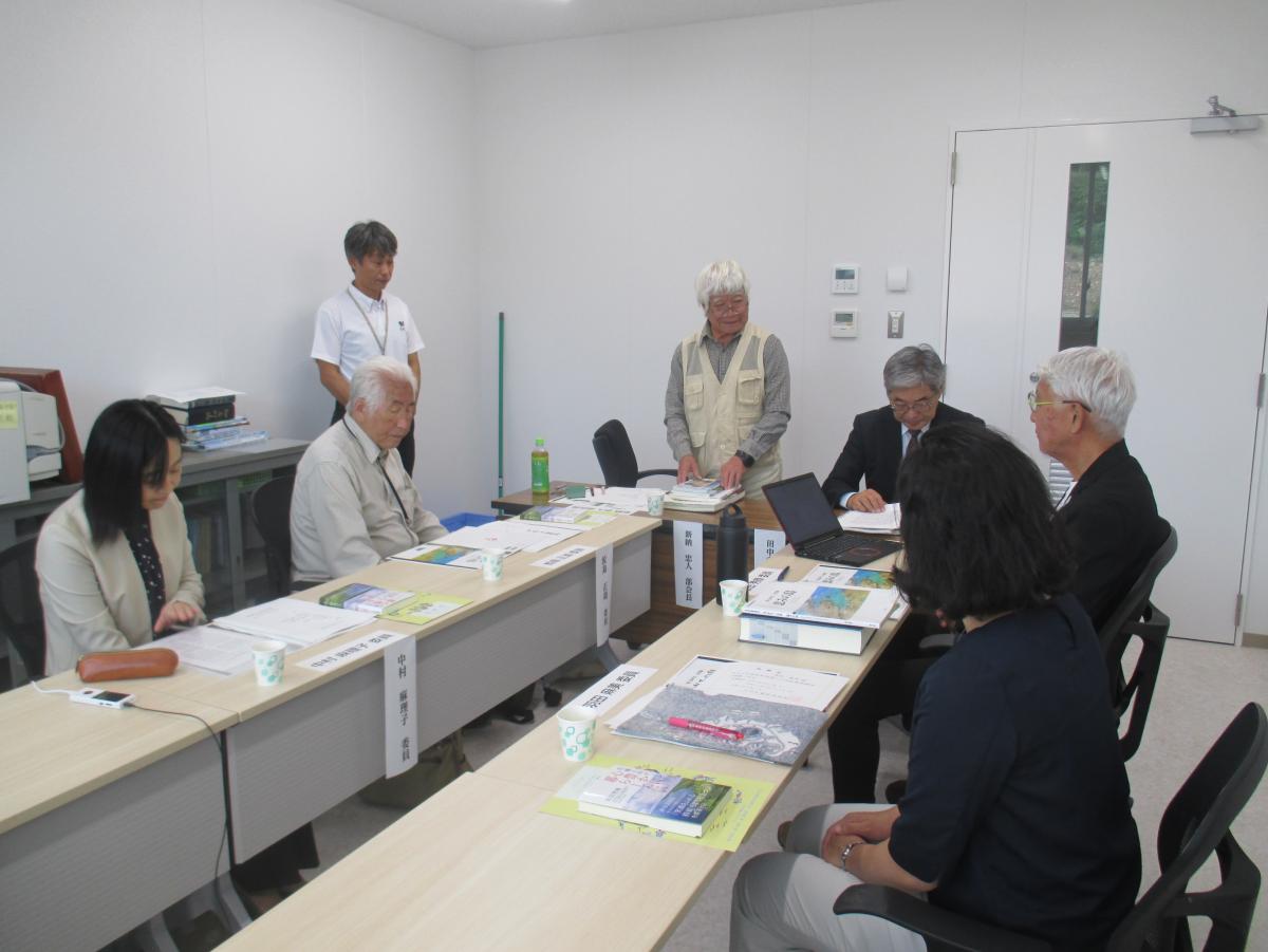 関係者達が向かい合うように長テーブルを囲み、中央では白いポロシャツの男性が立ち、机上には書類やノートパソコンが広げられ自然部会の様子の写真